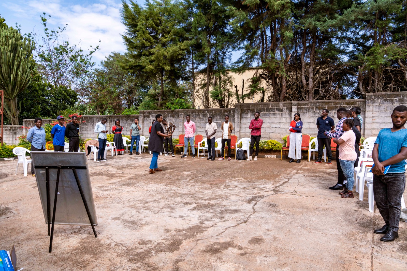 Community members gathered in a circle at an outdoor workshop venue, standing and engaging in dialogue. Concrete block walls and green trees create the background. The setting conveys unity, participation, and collaborative peacebuilding efforts.